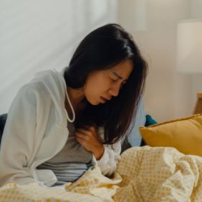 A woman with long black hair is sitting up in bed wincing in pain and one hand is on her chest.